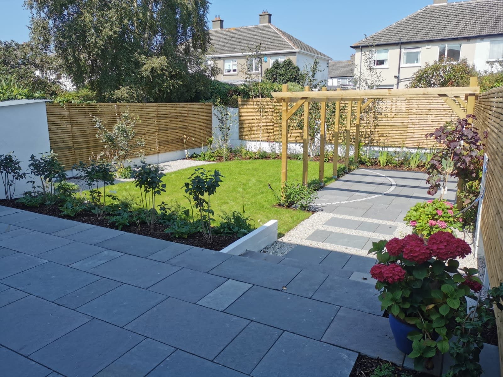 Finished garden with stone paving, a green lawn, planted borders, timber screening and a pergola