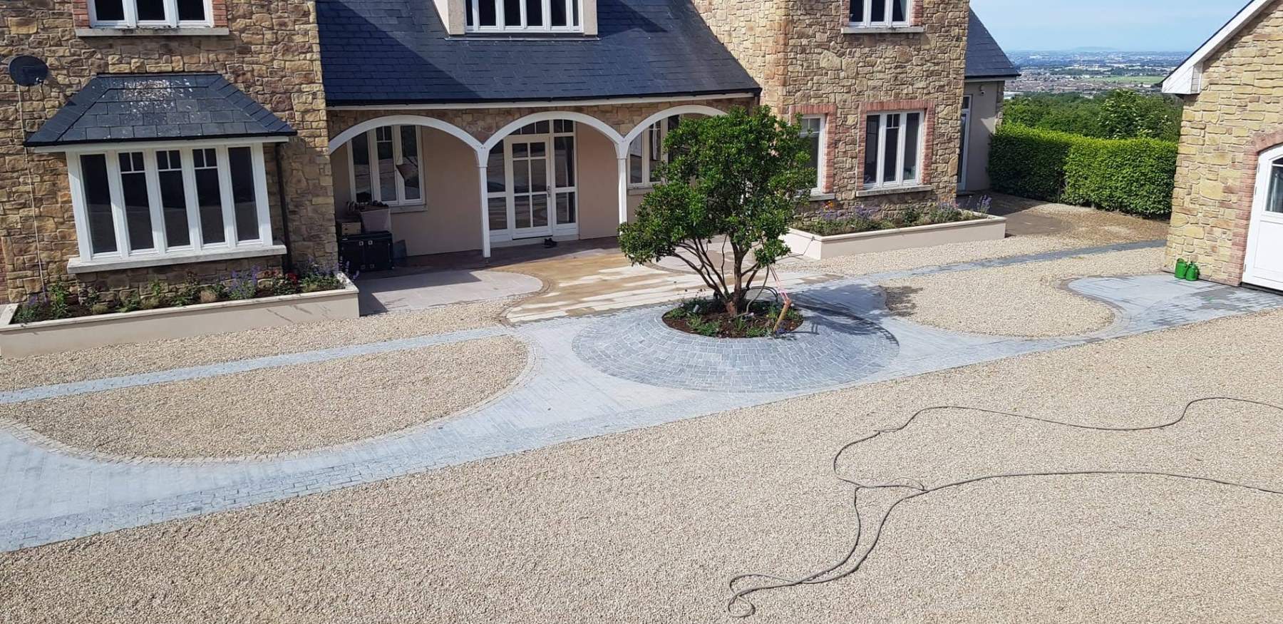 The same front approach after landscaping — pale gravel, curved block-paved walkways, a circular paved apron with an ornamental tree and planted beds